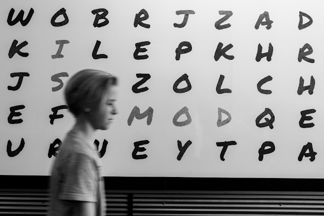 アルファベットが書かれたボードの前を通る、1人の少女のモノクロ画像