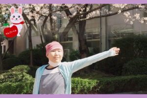 満開桜の木の下で左腕を広げ上げているピンク色帽子を被った女性がん患者