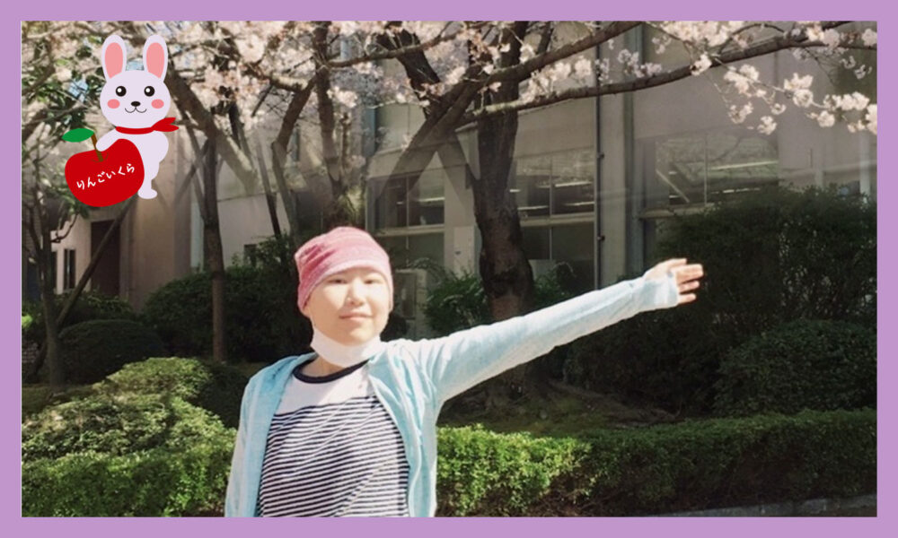 満開桜の木の下で左腕を広げ上げているピンク色帽子を被った女性がん患者