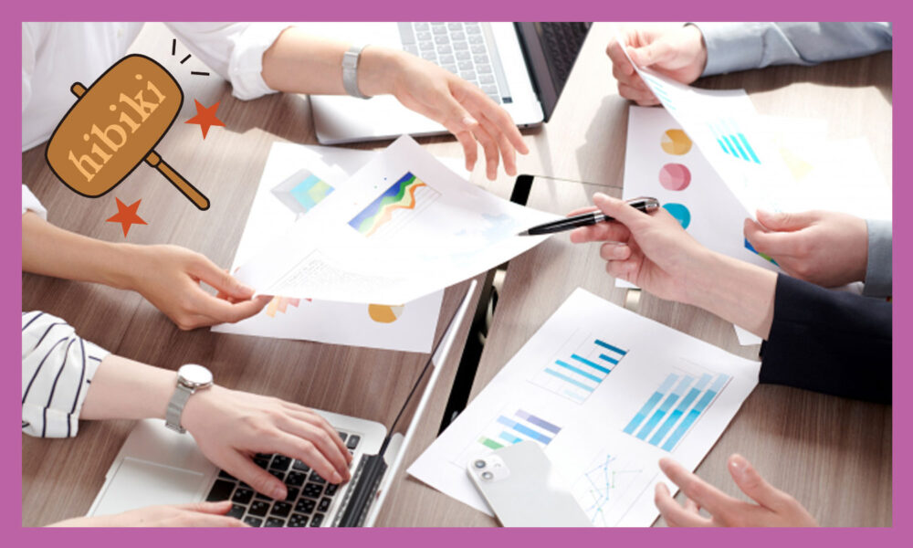 会議中で資料が散らばっている。４人の人の手だけが写っている様子う。