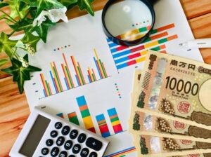 机の上に、グラフ用紙、虫眼鏡、お金、電卓が散らばっている。
