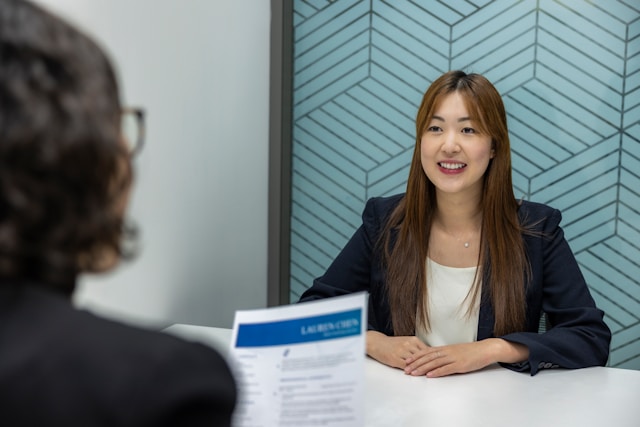書類を手に持っている眼鏡をかけたスーツ姿の女性と、その前に座っている髪の長い、スーツ姿の女性
