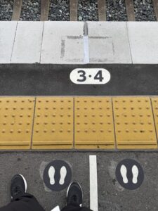 駅のホーム。電車の乗り場3・4と番号が書いてある。その手前に、点状ブロックと線がある点字ブロック一番手前には執筆者の足がある画面。