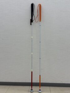 壁に立てかけられた2本の白杖。左が太い持ち手で太い白杖で右がオレンジのグリップで細い白杖