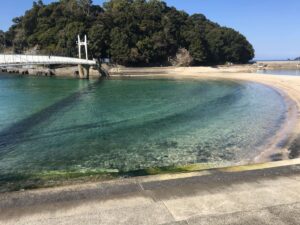 画面右側に砂浜と左側に続く海。奥に橋が見える。