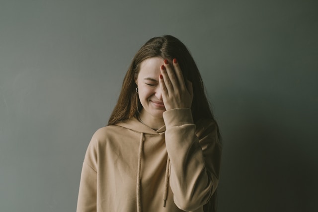 片手で顔を覆いながら目を閉じている、ベージュのパーカーを着た若い女性