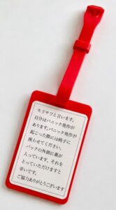 シリコン製の赤いヘルプマークの上に貼られたシール。このシールに「モリサワと言います。自分はパニック発作があります。パニック発作が起こった際には椅子に座らせてください。バックの外側に薬が入っています。それをとっていただけますと幸いです。ご協力ありがとうございます」と書いてある画面。