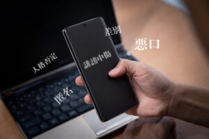 実物の写真として奥にパソコン、人の右手がスマートフォンを触っている。明朝体の白い文字で「悪口、差別、誹謗中傷、人格否定、匿名」と書いてある。