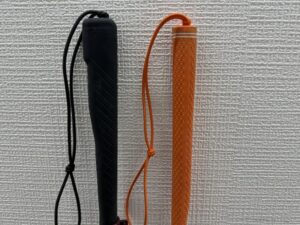 白杖のグリップ2種類の比較画像です。左側は、深い黒色で、しっかりと握れるように斜めに刻まれた溝が際立つグリップです。上部には持ち運びに便利な黒いゴム紐が取り付けられています。右側は、鮮やかなオレンジ色で、滑り止めとして表面全体に小さな凹凸(ディンプル加工)が均一に施されたグリップです。こちらも上部に、同じオレンジ色のゴム紐が取り付けられています。