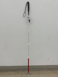 白杖を垂直に立てた全体の画像。上部の黒いカーボングリップから、白、白、白、そして先端(石突)に近い最後の段だけが赤色の反射テープに切り替わっている。杖は床に接しており、その最下部には、丸くて白い、パームチップ(かさの厚いキノコの形をした石突)が装着されている。白壁とタイル床の背景。