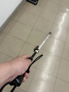 この画像は、実際に使用する際のグリップの持ち方と、杖の先が前方に伸びている様子を伝えています。
白杖を実際に手に持って使用しているシーンを、持ち主の視点(一人称視点)で捉えた画像。手前には黒いカーボングリップを握る人の左手が写っており、杖は白いタイル床の廊下に向かって前方に真っ直ぐ伸びている。杖の中ほどには茶色のクリップが、先端にはパームチップ(かさの厚いキノコ形)の石突が見える。