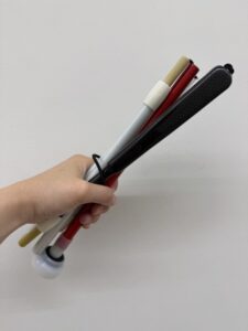 人の左手が、一束の折りたたみ式白杖(はくじょう)を斜めに持っている。束ねられているのは、上から順に:ベージュのシャフトの中身と白のシャフト、赤いシャフト、そして黒いカーボン調のシャフトとグリップを持つ白杖。各シャフトは折りたたまれており、黒いゴムバンドで束ねられている。赤いシャフトの白杖の先端(チップ)が一番下にきている。