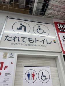 天井付近に設置された、「だれでもトイレ」を案内する大型の掲示板を写した写真です。上部には、大人用ベッドのピクトグラム（介助者が乳幼児にオムツを替えている様子）と、車椅子利用者（国際シンボルマーク）のピクトグラムが並んで掲示されています。その下に、日本語で「だれでもトイレ」と大きな文字で書かれ、その横に小さなレインボーフラッグの絵文字が添えられています。さらに「どなたでもご自由にお使いください」と説明文があります。掲示板の下には木目調のドアがあり、ドアには別の小さなサインが掲出されています。ここには、男女別のトイレのピクトグラム（左：男性・青色、右：女性・赤色）と、車椅子利用者のピクトグラムが並んで描かれています。壁面には他にも、1階フロアマップの一部や、「免税（Tax）」と書かれた赤い看板の一部が写っています。