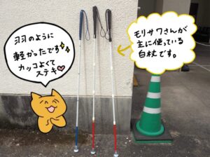 壁際に3本の白杖が並んで立てかけられている写真。青や黒の持ち手とストラップが付いており、それぞれ形やデザインが異なる。右側の吹き出しに「モリサワさんが主に使っている白杖です。」
左側には「羽のように軽かったです。 カッコよくてステキ。」と、中鮮のイラストと共にセリフがイラストで描かれている。