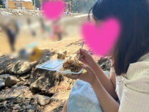 川沿いで焼きそばを頬張る女の人、顔はピンクのハートで隠されている