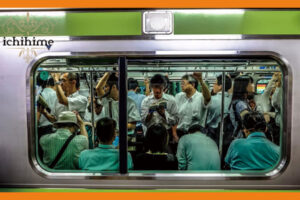 満員の電車の中で、立って本を読んでいる男性の画像